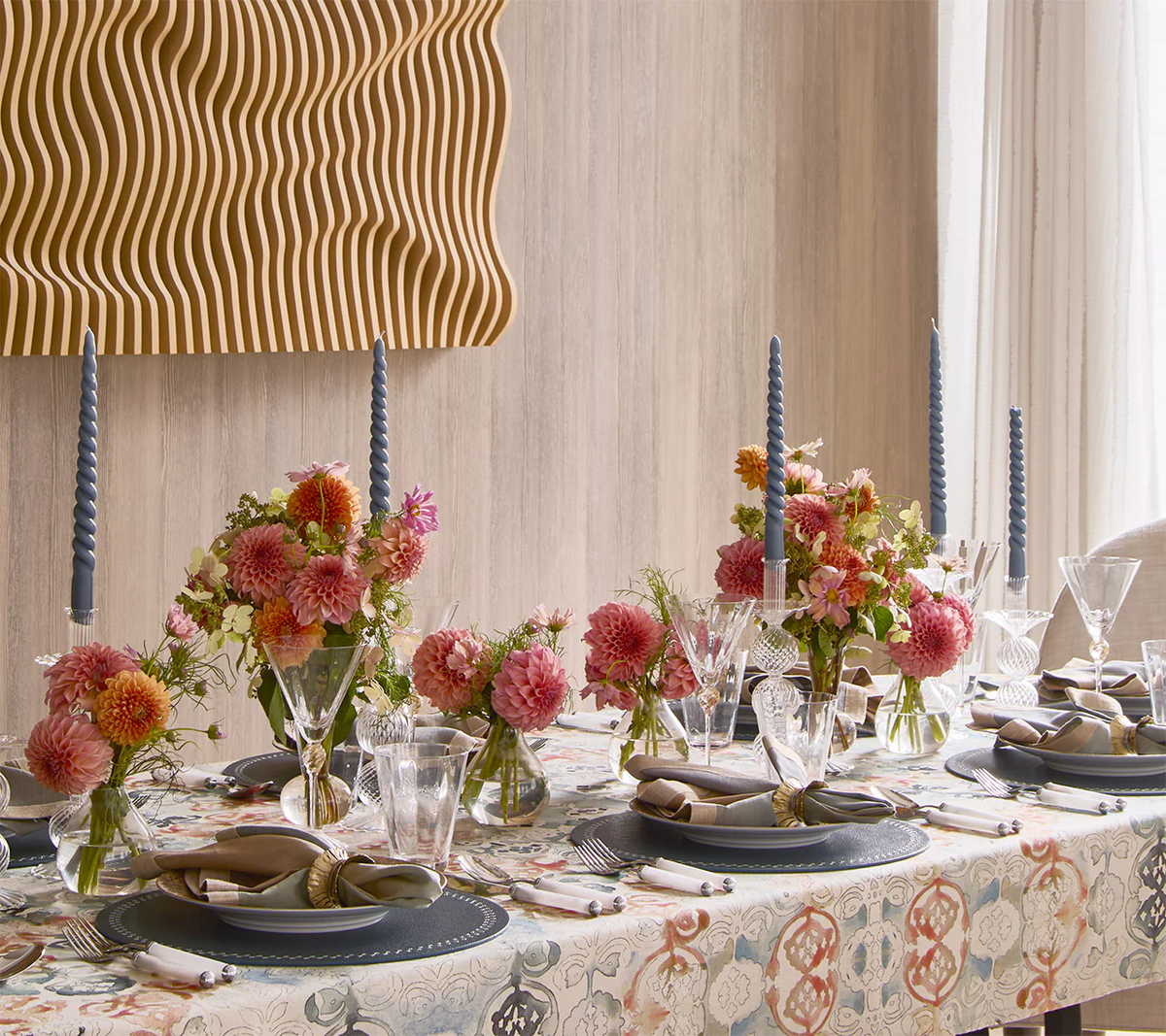 A dining table is elegantly set with pink and orange florals, blue twisted candles, glassware, and napkins finished with Kim Seybert’s Clinched Napkin Ring in Gold & Silver. A wavy wooden wall art piece hangs above the light wood wall.