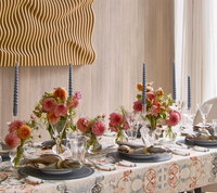 A dining table is elegantly set with pink and orange florals, blue twisted candles, glassware, and napkins finished with Kim Seybert’s Clinched Napkin Ring in Gold & Silver. A wavy wooden wall art piece hangs above the light wood wall.