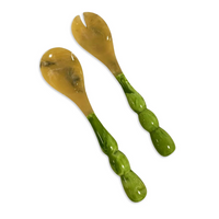 The Beatriz Ball Resin Rio Bubble Salad Servers feature translucent yellowish-brown heads and sculpted green bubble handles—one forked, one rounded—and are displayed on a white background.