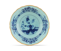 The Oriente Italiano Dinnerware Iris by Ginori 1735 is a porcelain plate with scalloped edges, a blue chinoiserie floral motif on a pale background, and a thin gold rim.