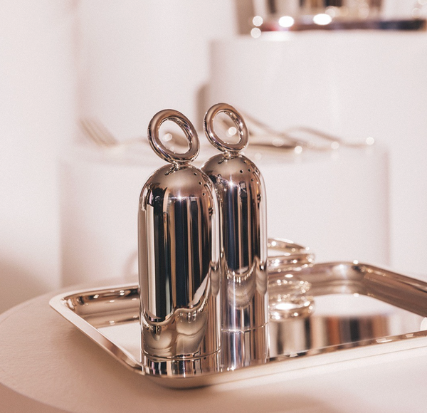 Two Christofle Vertigo Salt and Pepper Shakers by Christofle, featuring signature ring handles, sit on a reflective tray with blurred cutlery and dishes in the soft, neutral background.