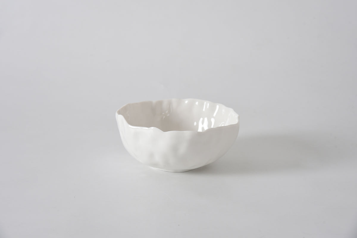 The PAMPA BAY Willow White Bowl, featuring a wavy rim and white ceramic finish, sits on a smooth light gray surface with a plain light gray background.