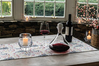 A handmade Simon Pearce Vintner Wine Decanter with a marble stopper, filled with red wine, stands on a floral table runner beside a glass, wine bottle, and candle on a wooden table, large windows and flowers in the background.