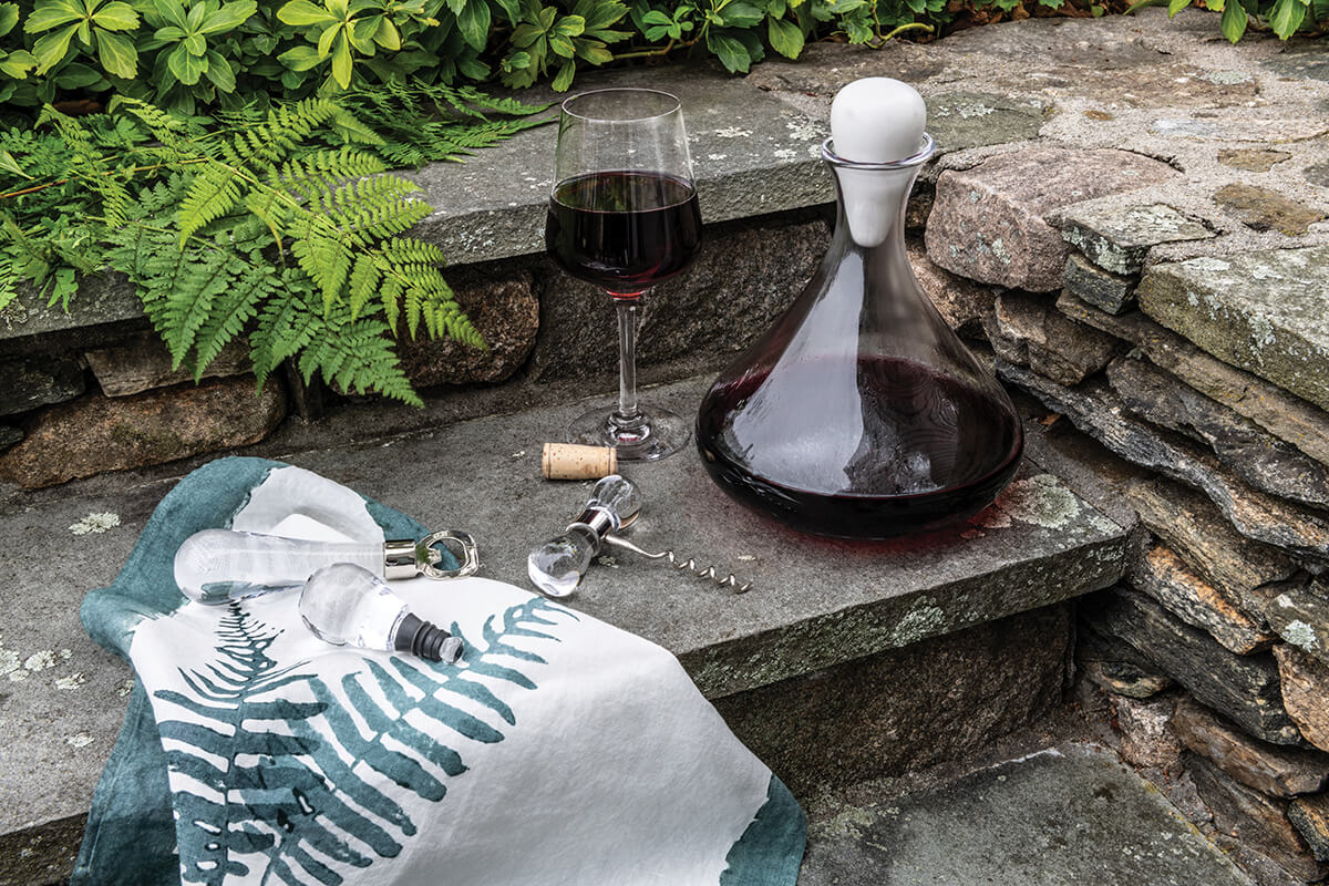 The Simon Pearce Vintner Wine Decanter With Marble Stopper, paired with a corkscrew, sits on stone steps beside a green fern-print cloth, surrounded by lush ferns and rustic stone walls.