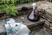 The Simon Pearce Vintner Wine Decanter With Marble Stopper, paired with a corkscrew, sits on stone steps beside a green fern-print cloth, surrounded by lush ferns and rustic stone walls.