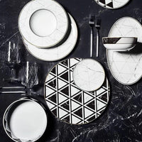A modern table setting showcasing Carrara by Coline Le Corréa dinnerware from Vista Alegre—porcelain plates, bowls, and glasses with bold black-and-white geometric and marble patterns—arranged on a dark Carrara marble surface.