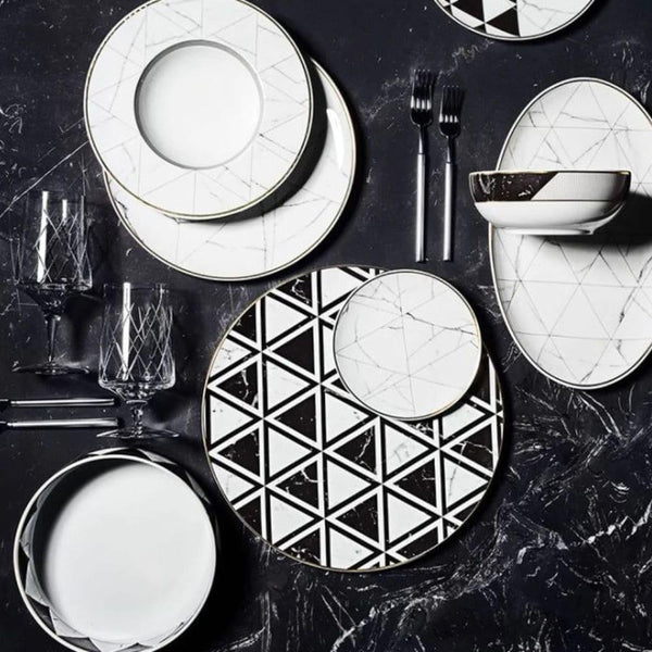 A modern table setting showcasing Carrara by Coline Le Corréa dinnerware from Vista Alegre—porcelain plates, bowls, and glasses with bold black-and-white geometric and marble patterns—arranged on a dark Carrara marble surface.