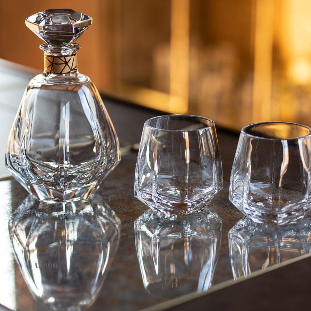The Vista Alegre Gemstone Highball Set of 2 features two modern tumblers beside a crystal decanter with a diamond-shaped stopper, all displayed on a reflective surface against a warm, blurred background.