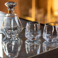 The Simon Pearce Vintner Wine Decanter With Marble Stopper and two matching glasses rest on a reflective surface, casting elegant reflections. The softly blurred background glows with warm, golden tones.