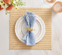 A Vista Reversible Placemat set in Natural & Brown by Kim Seybert holds a white plate with gold trim, a blue napkin with ruffled white edge in a gold ring; flowers and pink glassware are nearby.