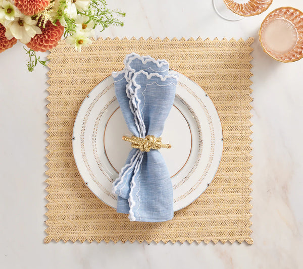 A Vista Reversible Placemat set in Natural & Brown by Kim Seybert holds a white plate with gold trim, a blue napkin with ruffled white edge in a gold ring; flowers and pink glassware are nearby.
