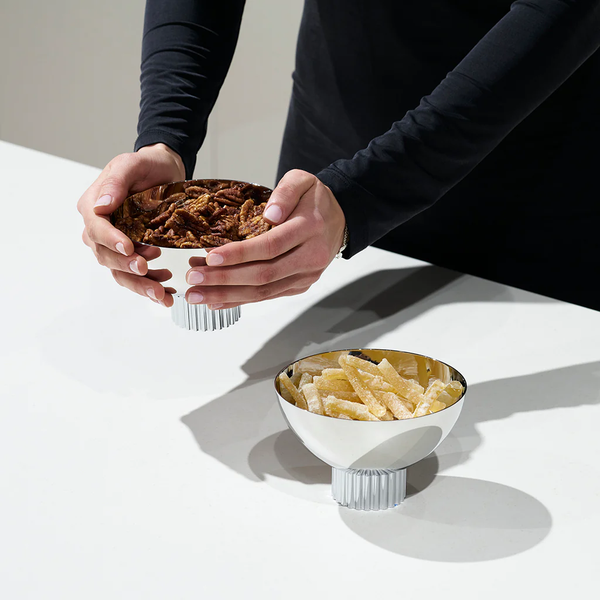 A person in a black long-sleeve shirt holds the Anna New York Striations Silver Nut Bowl filled with mixed nuts above a white counter, while another bowl of chips sits nearby.