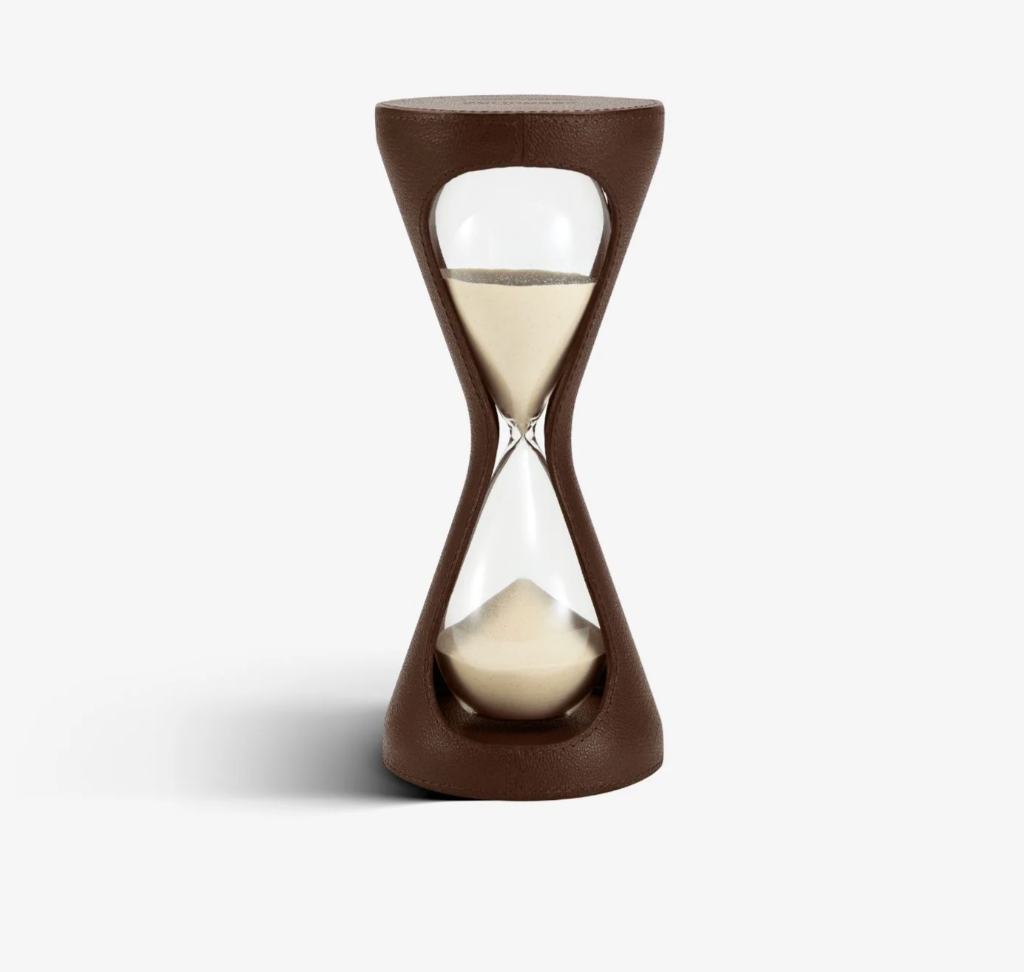 The Assouline Mirage Luxury Leather Hourglass features light sand and a sleek dark brown leather exterior, elegantly displayed on a white background—an ideal decorative timer for any space.