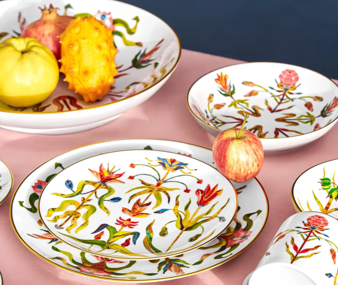 L’Objet Grand Tour Dinnerware in white porcelain with colorful floral patterns and 24k gold accents, displayed with a large serving bowl containing fruit and a fresh apple on a side plate.