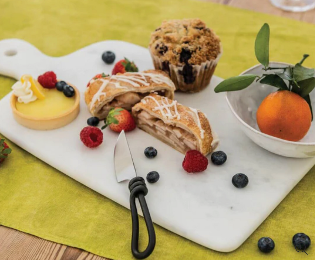 The Simon Pearce Marble Large Board displays a lemon tart, apple strudel slices, berries, a blueberry muffin, and clementine in a bowl on a green tablecloth. A small knife rests on the handcrafted marble surface.