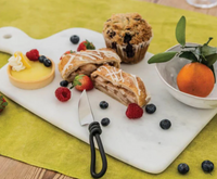 The Simon Pearce Marble Large Board displays a lemon tart, apple strudel slices, berries, a blueberry muffin, and clementine in a bowl on a green tablecloth. A small knife rests on the handcrafted marble surface.