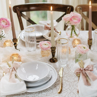 The Aria Dinnerware by Vietri features white stoneware plates, paired with glassware and gold cutlery. Ivory napkins with pink bows, pink flowers, and candles on a lace tablecloth complete this romantic table setting.