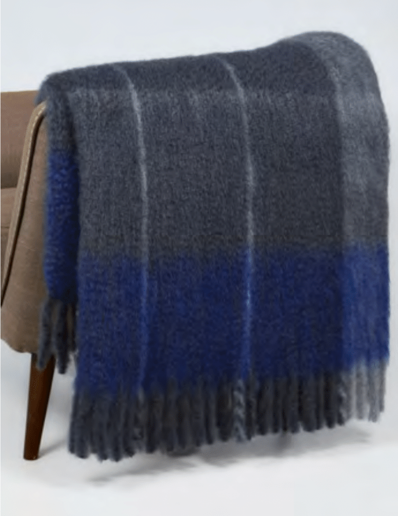 A soft, fuzzy Mantas Ezcaray Twist Frame Throw with wide gray and blue stripes and fringe is draped over a light brown chair; handcrafted in Spain.