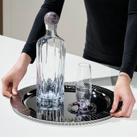 A person in a black shirt holds a round, reflective tray with a clear glass decanter and matching glass, both placed on Anna New York's Striations Silver Coasters Set of 4 in Holder, arranged on a white surface.