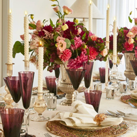Elegant dining table set with tall twisted candles, Kim Seybert Daphne Drinkware Set of 4 - Amethyst, gold-trimmed plates, ornate floral centerpieces in pink and burgundy, and linen napkins with gold rings for a festive touch.