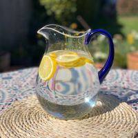 The Caspari Acrylic Handled Pitcher - Clear & Blue, filled with water and lemon slices, sits on a woven placemat atop a patterned outdoor table in sunny weather.
