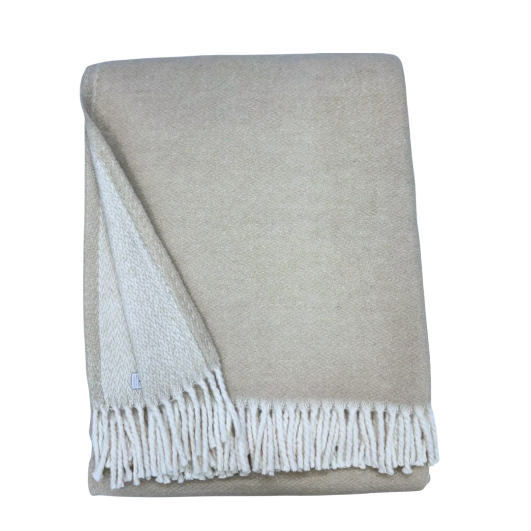 The Double Faced Solid Mini Chevron Throw by A Soft Idea is a beige, reversible blanket with white fringed edges. Displayed neatly folded on a plain white background, one corner reveals its lighter underside and fringe detail.