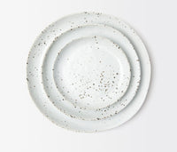 Four round, white ceramic plates with brown speckles from Blue Pheasant’s Marcus Dinnerware - White Salt Glaze set are stacked against a plain white background, highlighting their artisanal craftsmanship.
