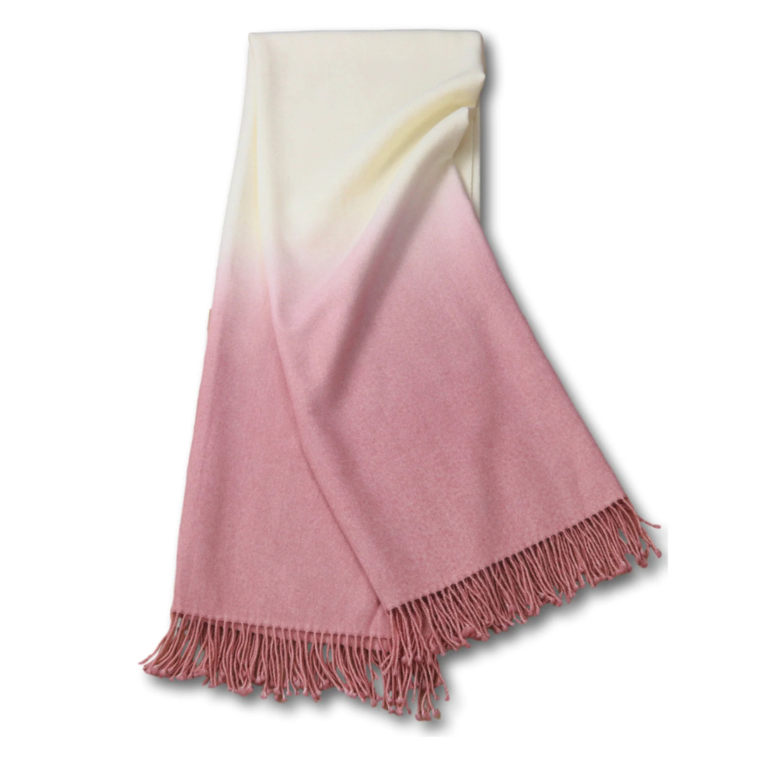 The Johanna Howard Dip Dye Throw is a soft woolen scarf with fringed edges, showcasing a gradient that gently fades from creamy white at the top to dusty rose pink at the bottom.