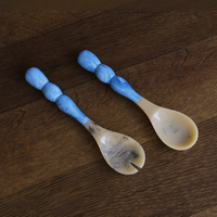 The Beatriz Ball Resin Rio Bubble Salad Servers feature marbled cream-colored spoon ends and blue bubble handles; one is a spoon, the other a matching fork, shown resting on a dark wooden surface.