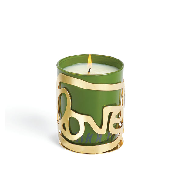 A green luxury candle from Amber Sakai, the Love Candle – Amour, sits in a gleaming 24K gold-plated holder with “love” elegantly shaped around it. The candle is lit, casting a warm glow against a plain white background.