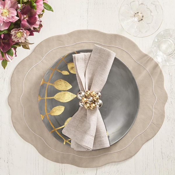 A table setting features the Kim Seybert Tailored Placemat Gray Set of 4, a gray plate with gold leaf design, a folded beige napkin in a decorative gold and silver napkin ring, plus a wine glass and pink flowers nearby.