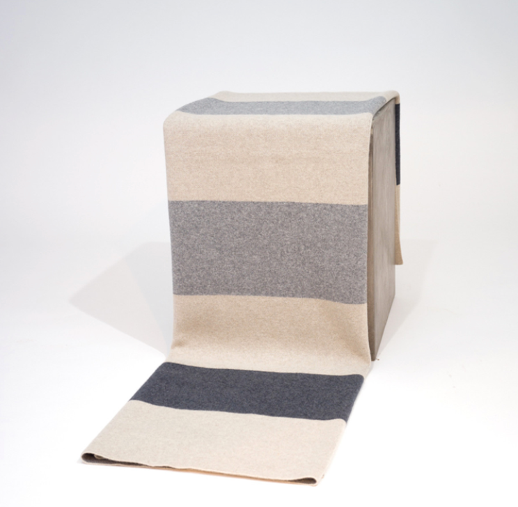 A Color Field Charcoal & Stone Grey Cashmere Throw by Hangai Mountain Textiles with alternating wide horizontal stripes of charcoal, light grey, and cream draped over a square block.