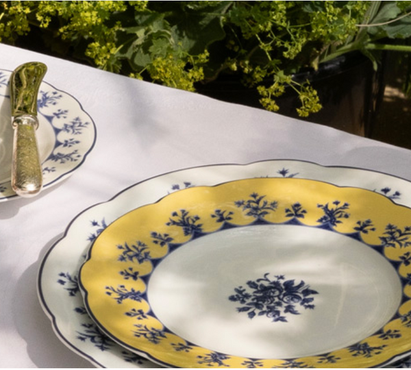 A close-up of stacked Bernardaud Chateaubriand Dinnerware plates featuring yellow and blue floral designs on a white tablecloth, with silver cutlery and green plants in the background. Sunlight casts soft shadows on the table.