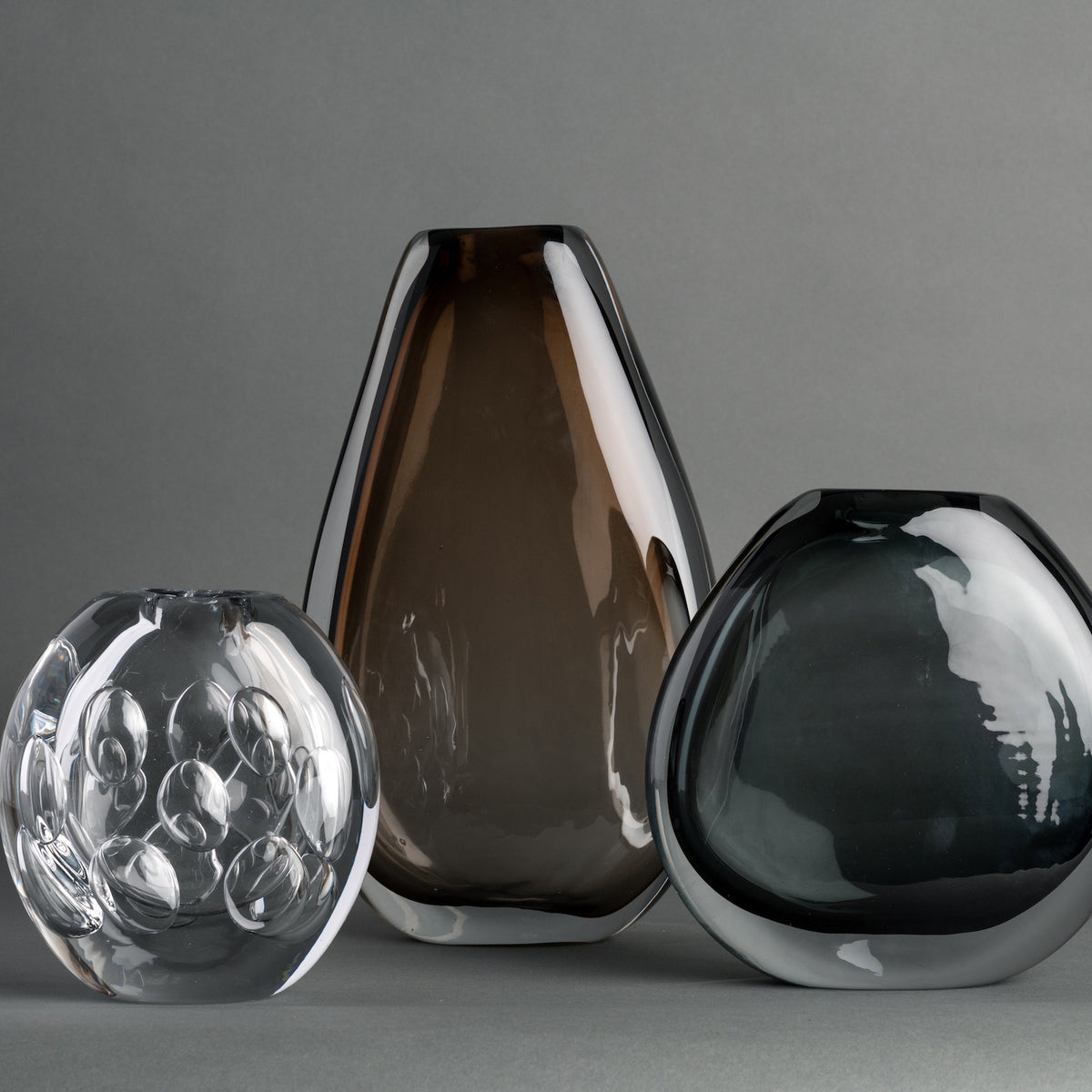 Three modern glass vases—a contemporary Whitney Vase by FlowDecor Inc. featuring bubble patterns in clear glass, one tall vase in smoky brown, and one rounded vase in dark gray—are arranged against a plain gray background.
