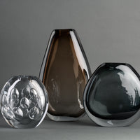 Three modern glass vases—a contemporary Whitney Vase by FlowDecor Inc. featuring bubble patterns in clear glass, one tall vase in smoky brown, and one rounded vase in dark gray—are arranged against a plain gray background.