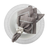 A gray linen napkin with lace trim, held by a silver floral napkin ring and placed on a white plate with a striped rim, rests atop the elegant Nomi K Shine Round White Placemat.