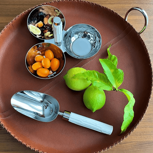 The Mary Jurek Tundra Triple Bowl With White Resin is displayed on a brown leather tray, featuring silver bowls with a hand-carved resin handle, metal scoop, green limes with leaves, nuts, dried fruit, kumquats, and an empty bowl.