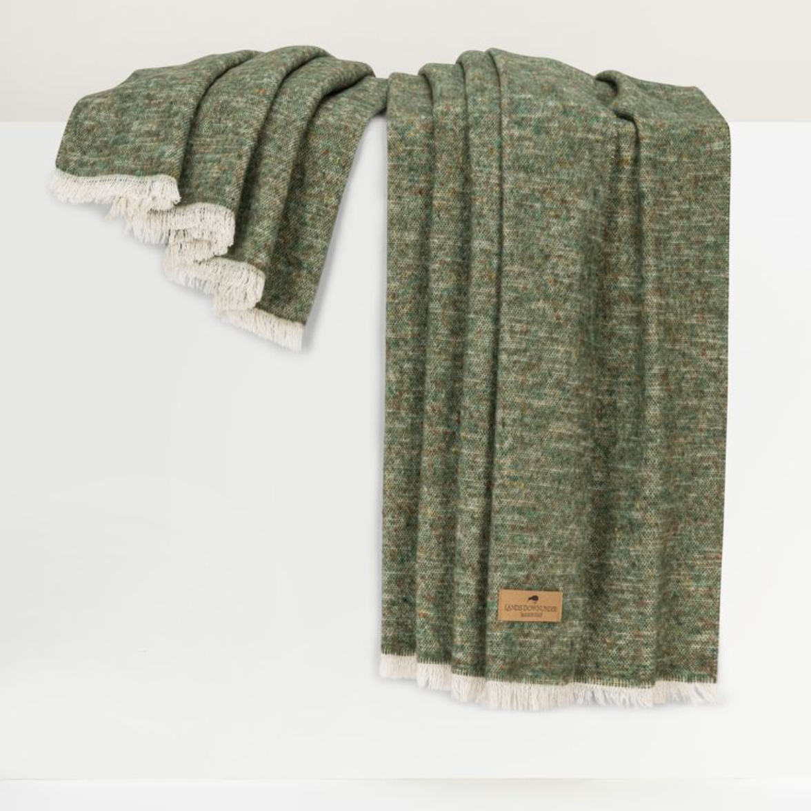 A Cinder Mist Throw by Lands Downunder, crafted in a modern style with green woven fabric and white fringe, is draped over a white surface. A brown rectangular label is attached to one corner of the blanket.