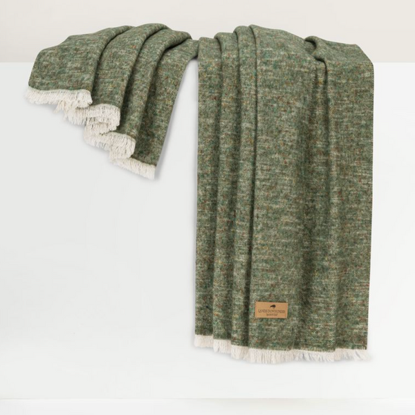 A Cinder Mist Throw by Lands Downunder, crafted in a modern style with green woven fabric and white fringe, is draped over a white surface. A brown rectangular label is attached to one corner of the blanket.