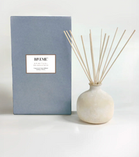The Blueme Spiritual Diffuser is a minimalist ceramic set with a round, pale vase and reed sticks, presented beside its light blue box. It offers therapeutic aroma wellness fragrances against a clean white background.