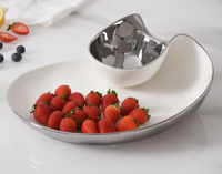 The PAMPA BAY Thin and Simple Chip & Dip, a white and silver porcelain two-section serving dish that's oven and dishwasher safe, holds fresh strawberries in the larger section, while the smaller one is empty. Blueberries and sliced strawberries sit on the countertop nearby.