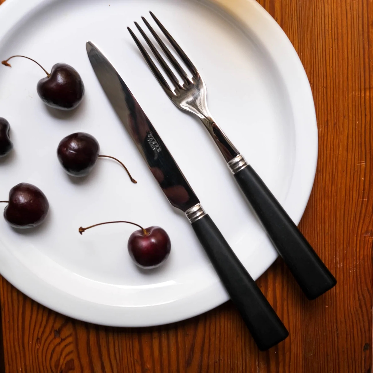 A white plate with five dark cherries sits beside black-handled Nature Flatware from Sabre’s 5 Piece Setting, elegantly arranged on a wooden table.