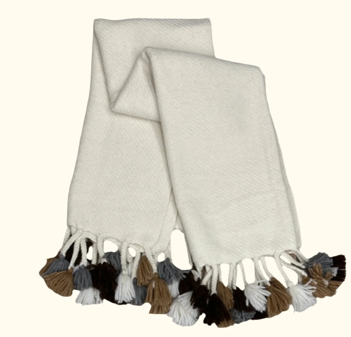 The Cusco Throw by Threads Soft Goods is a white woven alpaca scarf featuring brown, gray, black, and white tassels on both ends, shown on a light background.