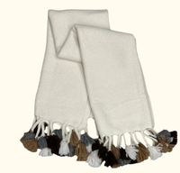 The Cusco Throw by Threads Soft Goods is a white woven alpaca scarf featuring brown, gray, black, and white tassels on both ends, shown on a light background.