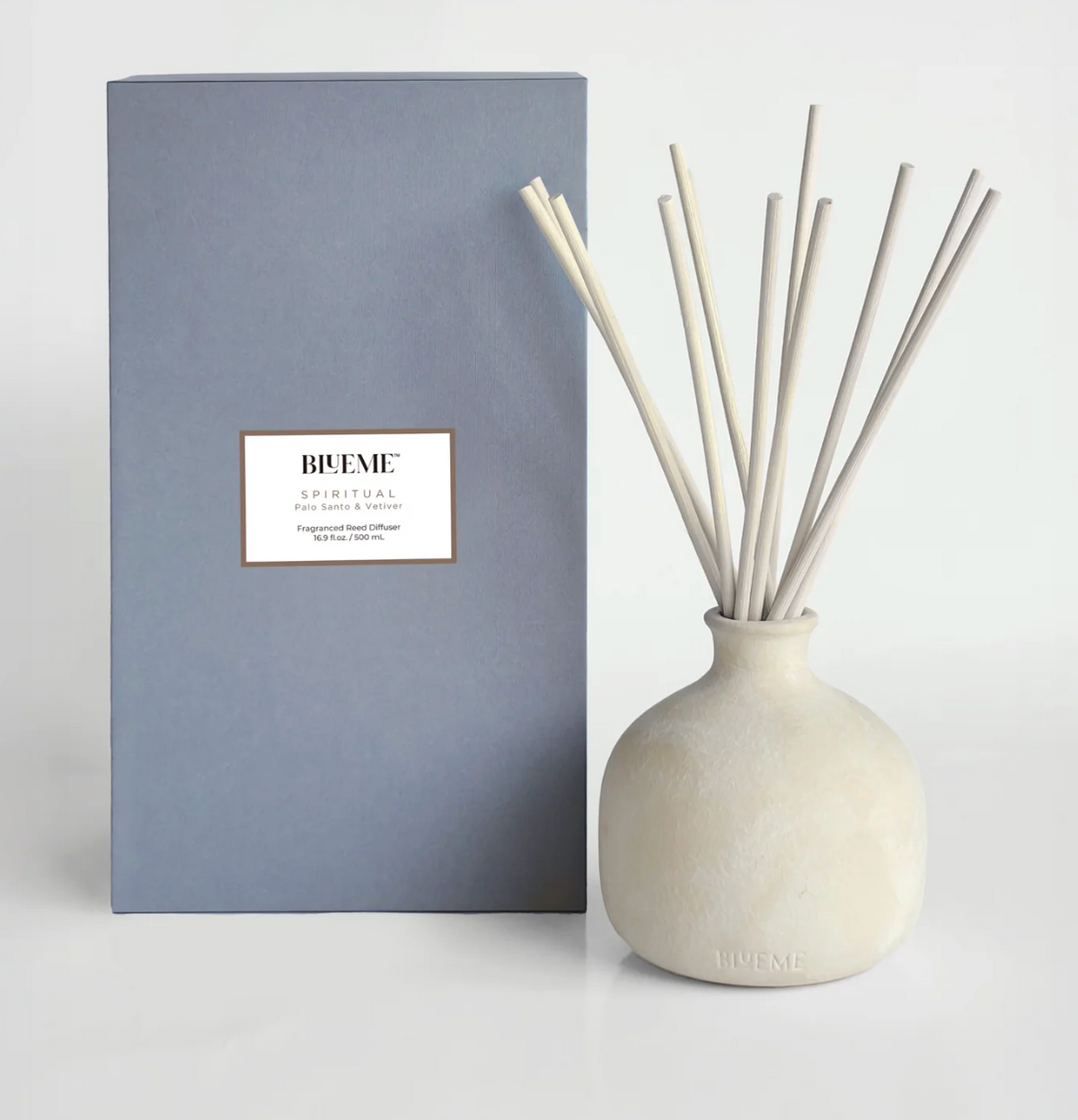 A beige ceramic Blueme Spiritual Diffuser with thin sticks stands by its tall gray box, filling the area with soothing, therapeutic wellness fragrances.