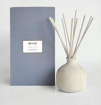 A beige ceramic Blueme Spiritual Diffuser with thin sticks stands by its tall gray box, filling the area with soothing, therapeutic wellness fragrances.