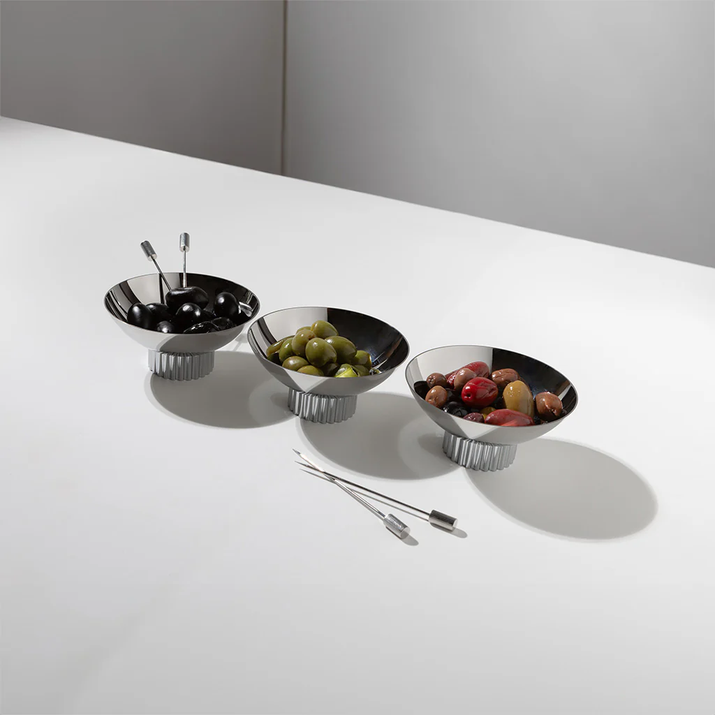 The Anna New York Striations Silver Cocktail Picks Set of 4 with holder is displayed in three silver bowls with assorted olives, stainless steel picks resting in one bowl and two on the white tabletop, against a minimalistic background.
