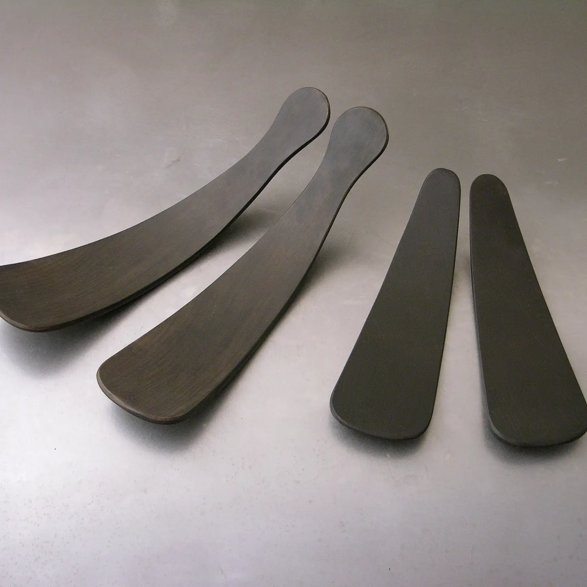 Four sleek, black Ebonized Oak Salad Tossers by Peterman—two long and two short—are neatly arranged on a gray surface, making a stylish dining accessory.