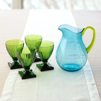 A Caspari Acrylic Handled Pitcher in turquoise & green sits beside four green goblets on a white table runner atop a light table with a shatter-resistant surface and acrylic dinnerware.