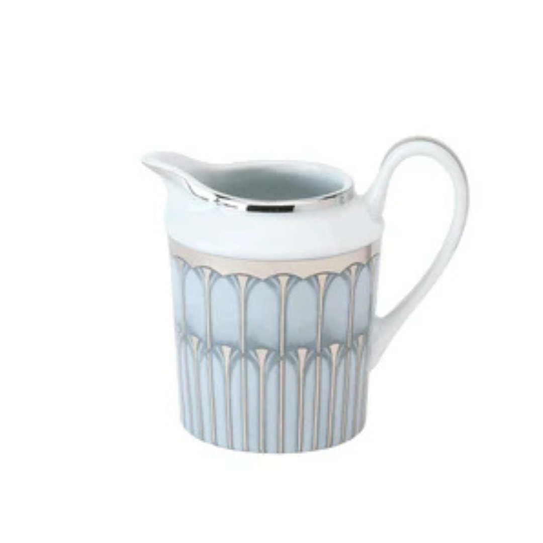 A white Limoges porcelain pitcher from Philippe Deshoulieres BIA's Arcades Platinum Dinnerware features a silver rim and handle with blue and silver geometric patterns around the body.